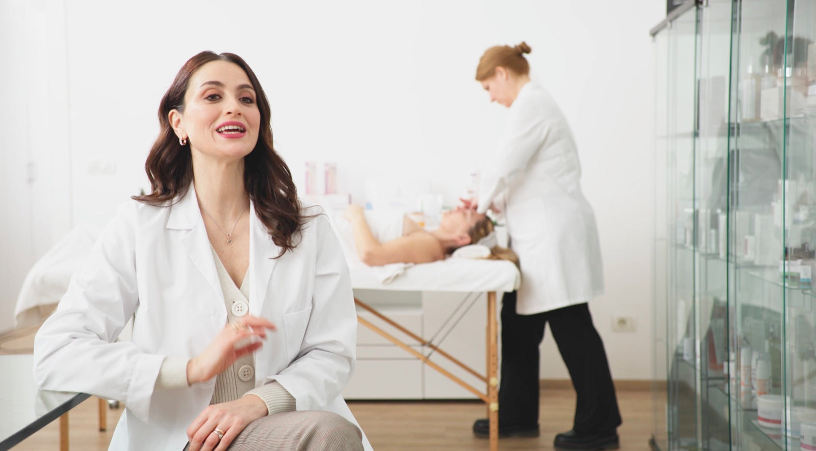 Un'esperta in camice in una cabina estetica; alle sue spalle un'estetista esegue un trattamento viso a una cliente sul lettino, accanto a una vetrina di prodotti.