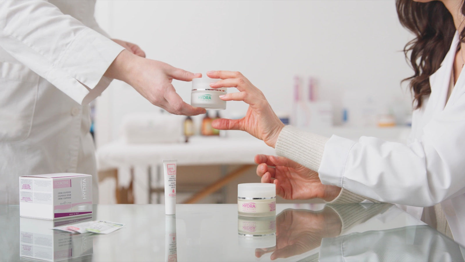 Due mani si passano un vasetto di crema Hydra Comfort sopra un banco in vetro con altri prodotti.