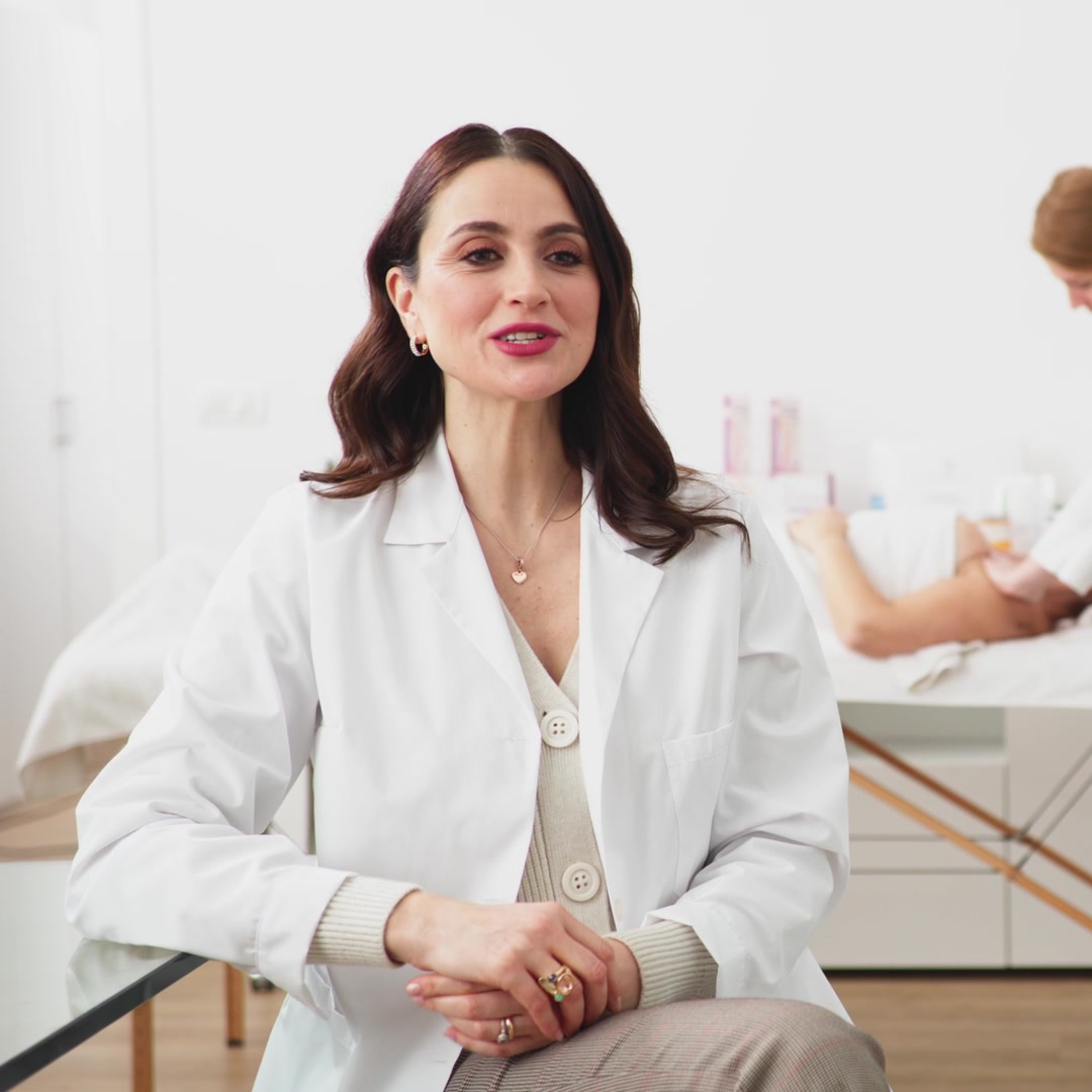 Un'esperta in camice bianco seduta in cabina; alle sue spalle un'estetista esegue un trattamento corpo.